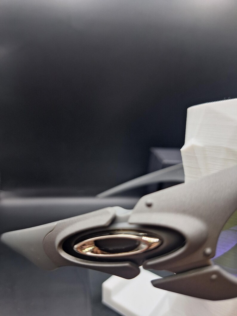 Op de afbeelding: Close-up van een grijs Oakley zonnebrilmontuur met een metallic gouden lensdetail. Het montuur heeft een strak, modern design met een prominent logo. De achtergrond is donker en onscherp.