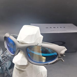 May include: Grey sunglasses with blue lenses are displayed on a white bust. The sunglasses have a sporty design with a matte finish. A black box with the brand name is visible in the background.