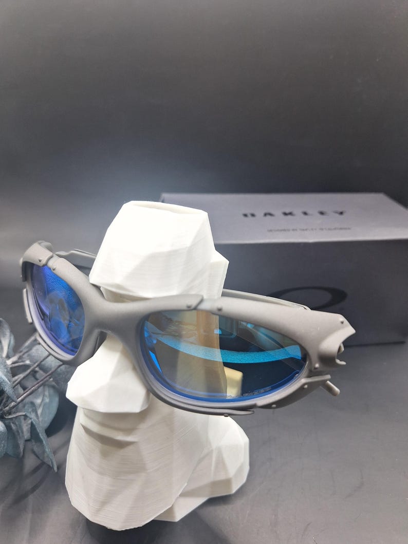 May include: Grey sunglasses with blue lenses are displayed on a white bust. The sunglasses have a sporty design with a matte finish. A black box with the brand name is visible in the background.