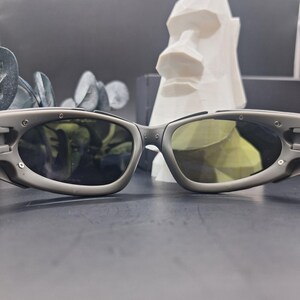 Może przedstawiać: Szare okulary przeciwsłoneczne z owalnymi soczewkami i ciemnym odcieniem. Okulary przeciwsłoneczne mają sportowy design z matowym wykończeniem. Biała geometryczna rzeźba głowy znajduje się w tle.