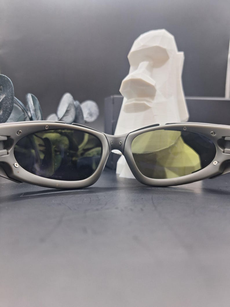 Może przedstawiać: Szare okulary przeciwsłoneczne z owalnymi soczewkami i ciemnym odcieniem. Okulary przeciwsłoneczne mają sportowy design z matowym wykończeniem. Biała geometryczna rzeźba głowy znajduje się w tle.