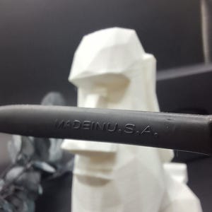 May include: A black object with the text "MADE IN U.S.A." is in front of a white, geometric sculpture of a head. The sculpture has a flat top and a defined nose and mouth. The background is dark.