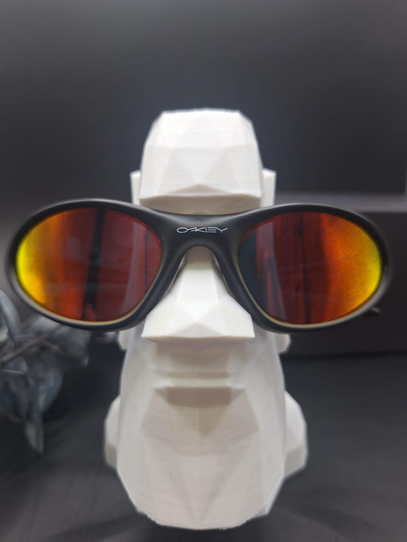 May include: Black Oakley sunglasses with orange and yellow lenses rest on a white geometric sculpture. The sunglasses have the Oakley logo in white on the frame. The sculpture is a stylized face.