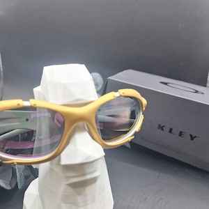 May include: Mustard-colored sunglasses with reflective lenses rest on a white, geometric sculpture. The sunglasses have a sporty design with a yellow frame and are positioned in front of a dark gray box with the brand name "OAKLEY" printed on it.