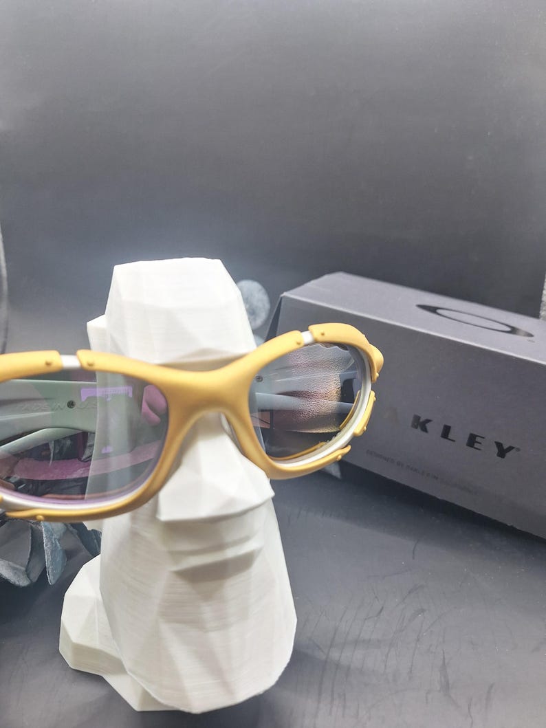 May include: Mustard-colored sunglasses with reflective lenses rest on a white, geometric sculpture. The sunglasses have a sporty design with a yellow frame and are positioned in front of a dark gray box with the brand name "OAKLEY" printed on it.