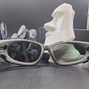 May include: Gray sunglasses with dark lenses. The sunglasses have a rounded rectangular shape and a thick frame. A white sculpture of a Moai head is in the background. The sunglasses are on a dark surface.