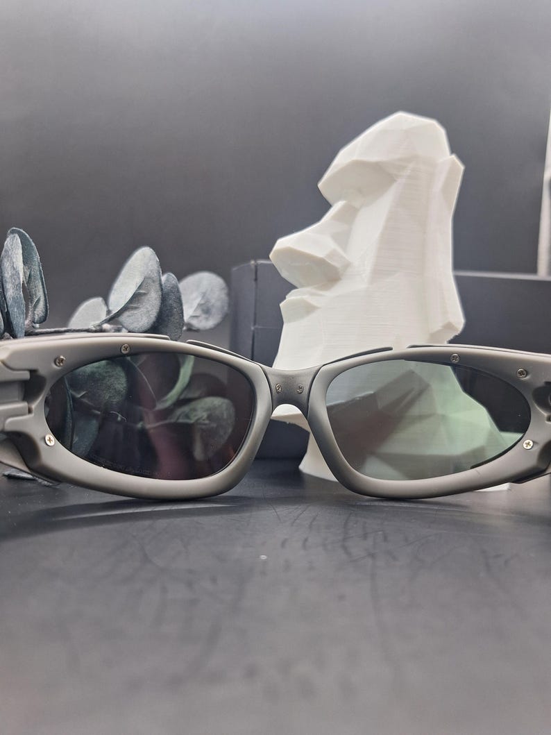May include: Gray sunglasses with dark lenses. The sunglasses have a rounded rectangular shape and a thick frame. A white sculpture of a Moai head is in the background. The sunglasses are on a dark surface.