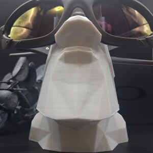 May include: Black sunglasses with yellow-tinted lenses rest on a white geometric display stand. The sunglasses have a dark frame and are positioned to showcase their design. The stand has a faceted, angular design.