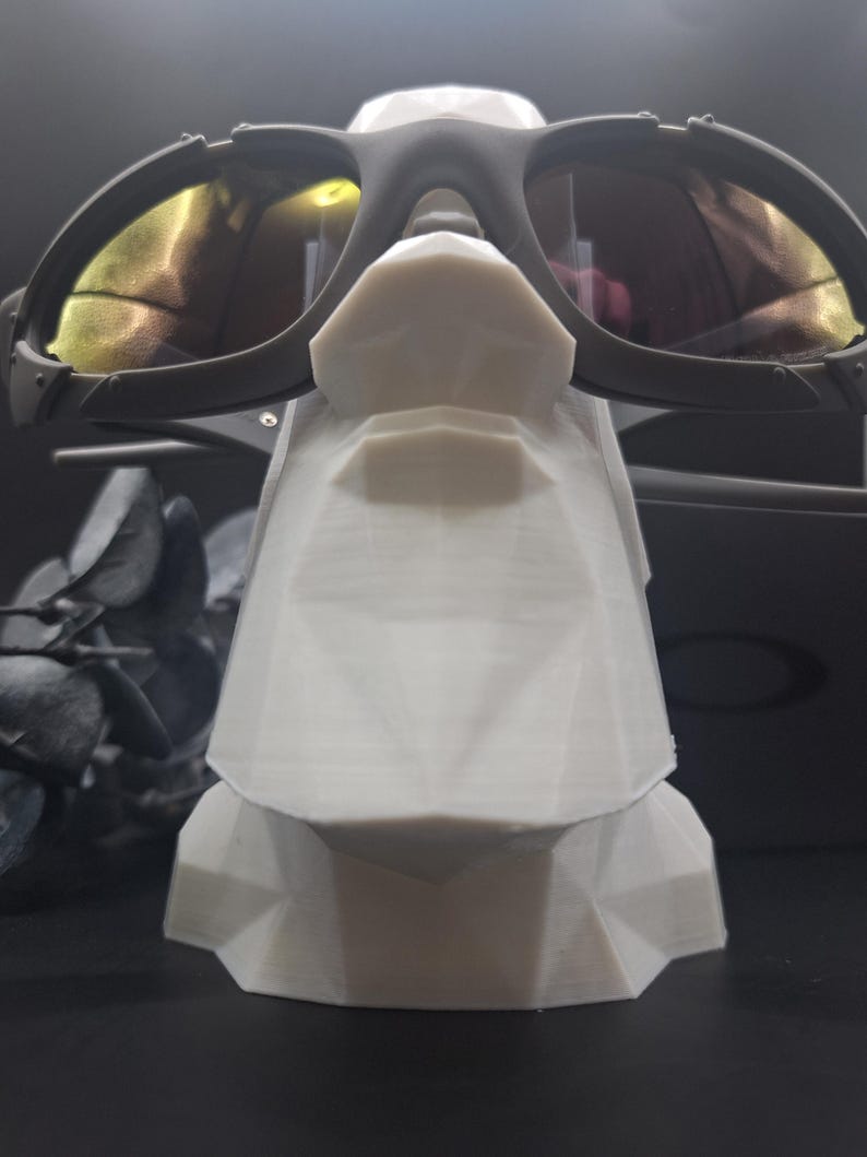 May include: Black sunglasses with yellow-tinted lenses rest on a white geometric display stand. The sunglasses have a dark frame and are positioned to showcase their design. The stand has a faceted, angular design.