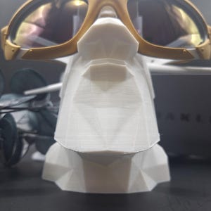 May include: Gold-framed sunglasses with reflective lenses rest on a white geometric display stand. The sunglasses have a sporty design with a gold bridge and temples. The stand is a modern, angular shape.