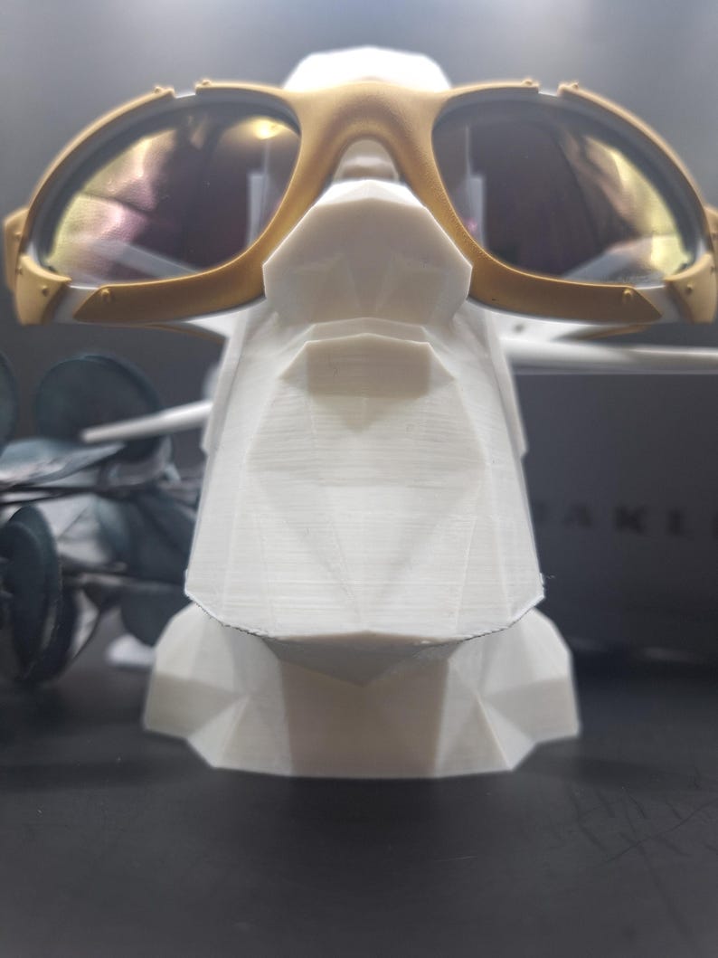 May include: Gold-framed sunglasses with reflective lenses rest on a white geometric display stand. The sunglasses have a sporty design with a gold bridge and temples. The stand is a modern, angular shape.