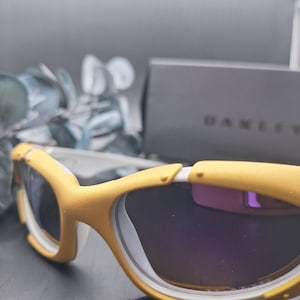 May include: Yellow and white sports sunglasses with dark lenses. The sunglasses have a curved design and are set against a blurred background with a box that says "OAKLEY".