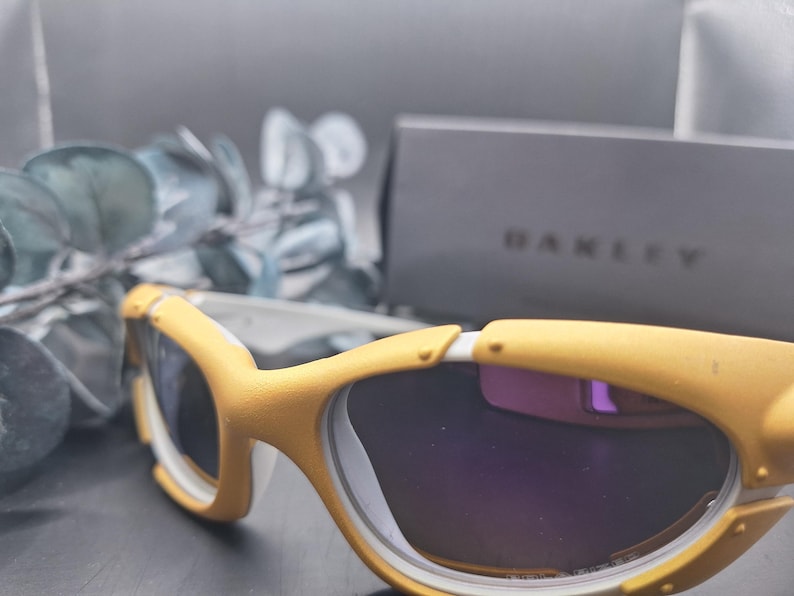 May include: Yellow and white sports sunglasses with dark lenses. The sunglasses have a curved design and are set against a blurred background with a box that says "OAKLEY".