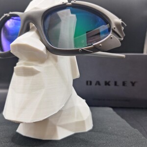 Op de afbeelding: Donkergrijze Oakley zonnebril met blauwgroene glazen rust op een witte geometrische sculptuur. De zonnebril heeft een kenmerkend, hoekig ontwerp. Een zwarte doos met de merknaam "OAKLEY" is zichtbaar op de achtergrond.