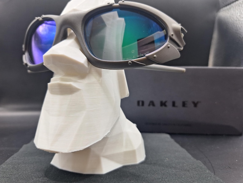 Op de afbeelding: Donkergrijze Oakley zonnebril met blauwgroene glazen rust op een witte geometrische sculptuur. De zonnebril heeft een kenmerkend, hoekig ontwerp. Een zwarte doos met de merknaam "OAKLEY" is zichtbaar op de achtergrond.