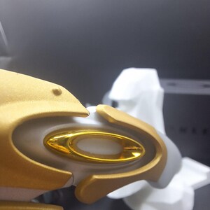 May include: Close-up of a gold and white object with a gold oval-shaped detail. The object has a smooth, metallic finish and appears to be part of a larger structure. The background is dark and out of focus.