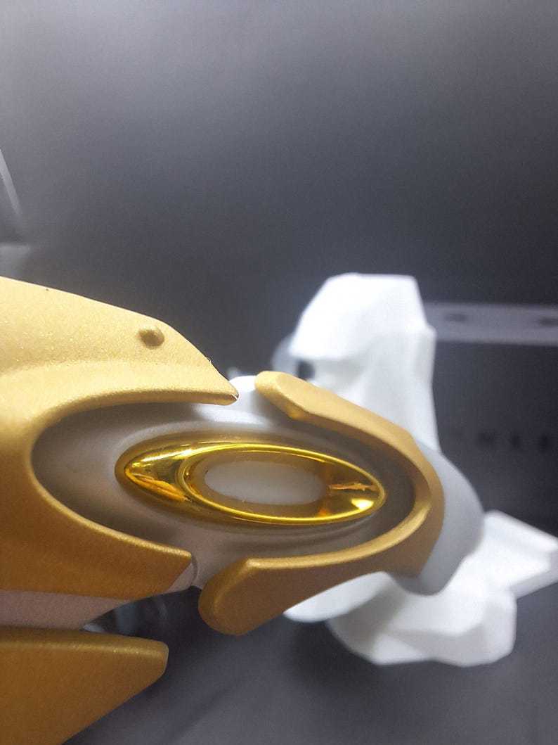 May include: Close-up of a gold and white object with a gold oval-shaped detail. The object has a smooth, metallic finish and appears to be part of a larger structure. The background is dark and out of focus.