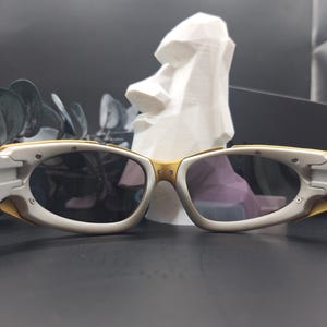 May include: White and gold-framed sunglasses with dark lenses. The sunglasses have a sporty design with a curved shape. The background includes a white sculpture of a Moai head and dark green leaves.