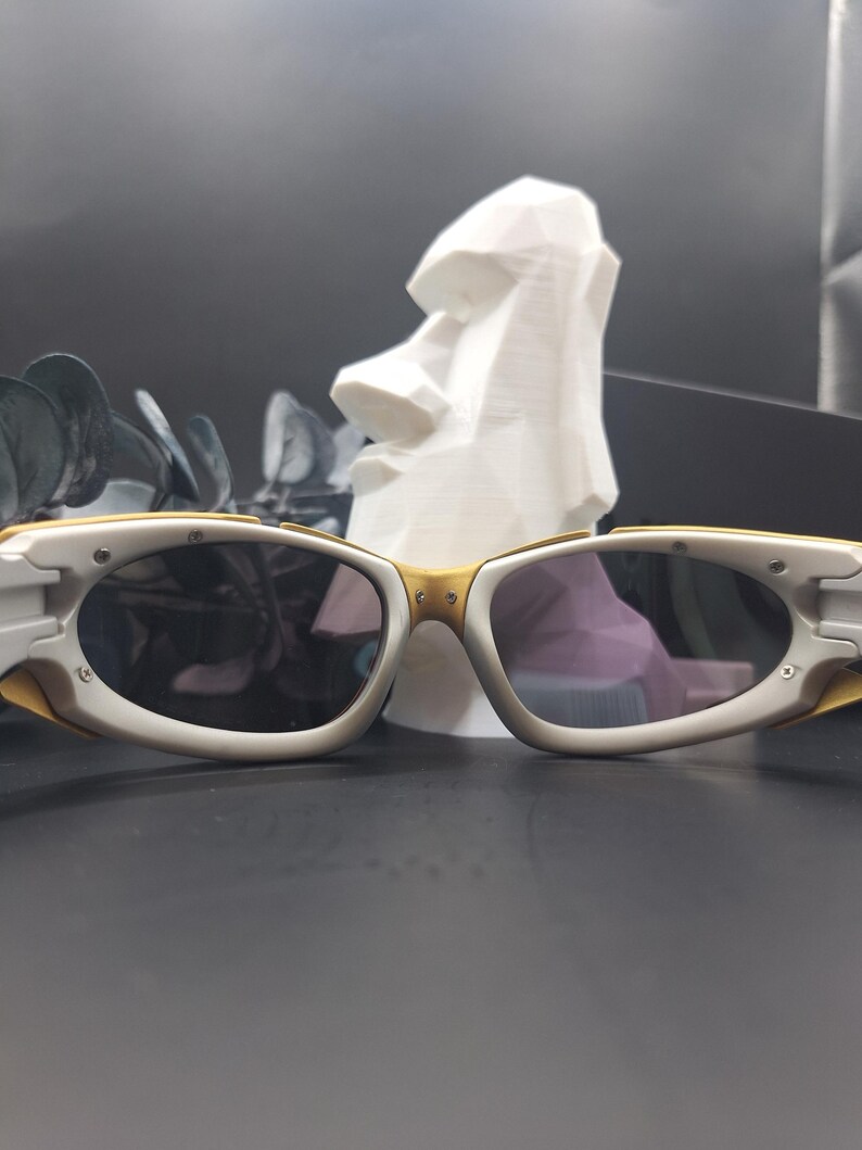 May include: White and gold-framed sunglasses with dark lenses. The sunglasses have a sporty design with a curved shape. The background includes a white sculpture of a Moai head and dark green leaves.