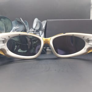May include: White and gold-toned sunglasses with oval lenses. The sunglasses have a white frame with gold accents on the bridge and temples. The lenses are dark, and the overall design is sleek and modern.