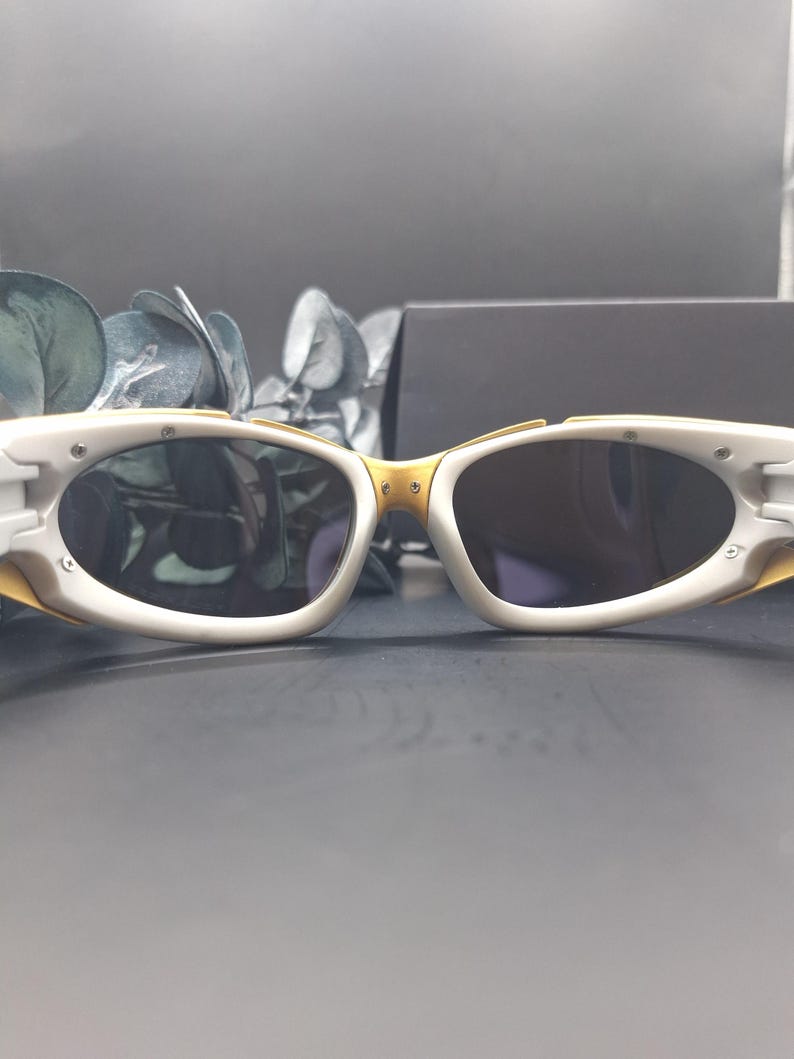 May include: White and gold-toned sunglasses with oval lenses. The sunglasses have a white frame with gold accents on the bridge and temples. The lenses are dark, and the overall design is sleek and modern.