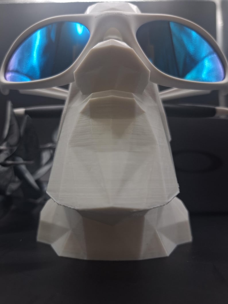 May include: A white, geometric sunglasses display stand holding a pair of sunglasses with blue reflective lenses. The stand has a stylized, angular design. The sunglasses are white with a modern frame.