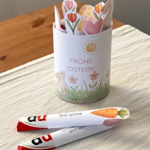 Puede incluir: Un recipiente cilíndrico blanco con las palabras "FROHE OSTERN" y una ilustración de un conejito. El recipiente contiene recortes de papel de flores, corazones y un conejito. Dos barras de chocolate envueltas están en primer plano.