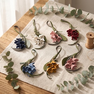 Puede incluir: Siete adornos florales de cuero decorativos con lazos para colgar. Cada pieza consta de un tallo y hojas de cuero verde combinados con pétalos multicolores en tonos azul oscuro, blanco, rosa claro, rojo intenso, azul claro, amarillo y malva.