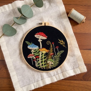 May include: A hand-stitched embroidery hoop featuring colourful mushrooms and ferns on a black background. The mushrooms have red, blue, and yellow caps with white spots. The embroidery is set in a wooden hoop and rests on a white linen cloth.