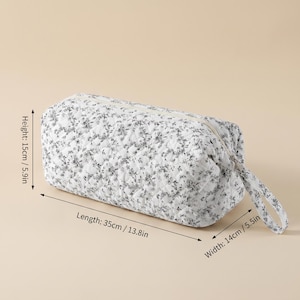 May include: A white quilted pouch with a black floral pattern, featuring a zip closure and a side handle. The pouch measures 15 cm in height, 35 cm in length, and 14 cm in width as indicated by the visible dimension labels on the product image.