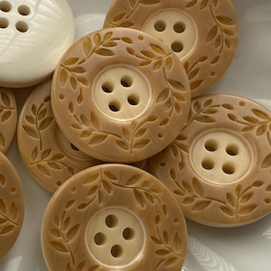 May include: A collection of beige and cream-colored buttons. The round buttons have four holes and are decorated with a leaf pattern around the edges. One large, round, cream-colored button is also present.