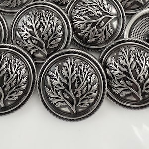 May include: A collection of silver-toned metal buttons with a tree design. Each button features a detailed tree motif within a circular frame, surrounded by a twisted rope-like border. The buttons are arranged on a white surface.