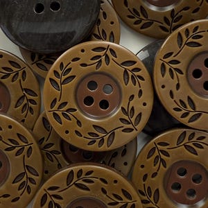 May include: A collection of decorative buttons in various shades of brown. The buttons have a circular shape with four holes and a leaf design around the edges. One button is dark gray.