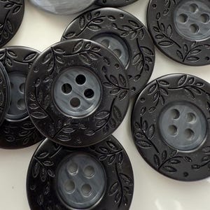 May include: A collection of round buttons in various sizes. The buttons are primarily black with a leaf design around the edges. One button is a light gray color. Each button has four holes for sewing.
