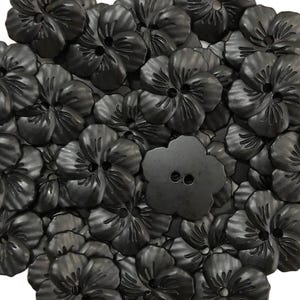 May include: A collection of matte black buttons shaped like five-petaled flowers. Each button features a central two-hole design and embossed radial lines on the petals, creating a textured surface. The buttons are arranged in a dense cluster.