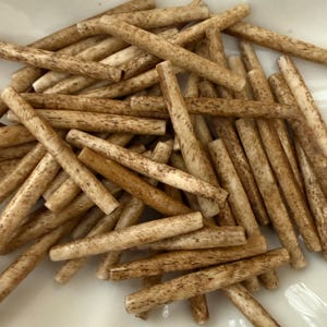 May include: A collection of small, cylindrical, light brown objects with darker brown markings, arranged in a white, scalloped dish. The objects appear to be made of a natural material, possibly wood or bone, and are scattered across the dish.