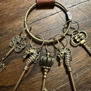 May include: A collection of antique-style keys in various designs, including seahorses and crowns, attached to a large brass ring and a brown leather strap. The keys are made of a bronze-colored metal.