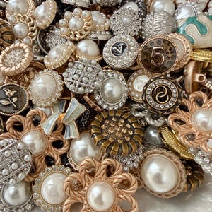 May include: A collection of assorted decorative buttons featuring various designs, including faux pearls, metallic finishes, intricate floral patterns, and geometric shapes. Some buttons display numbers or letters, while others are adorned with small clear crystals.