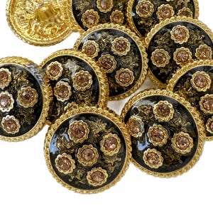 May include: A collection of decorative buttons with a gold-tone finish. The buttons have a black center with a floral design and small, light brown gemstones. The edges are trimmed with a gold rope-like design.