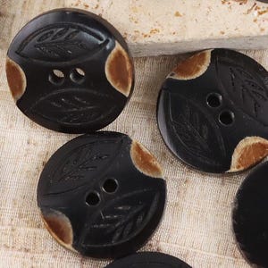 May include: A collection of round buttons in black and brown. Some buttons are solid black with two center holes, while others feature a black surface with two carved leaf patterns and brown accents on the edges, each with two center holes.