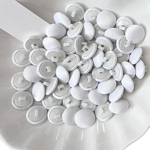 May include: Two white seashell-shaped dishes filled with white fabric-covered buttons. One dish has a label that says "Ivory". The buttons are small, round, and have a shank on the back. The buttons are likely for sewing or crafts.