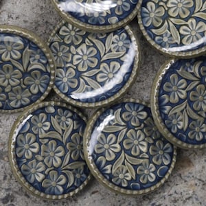 May include: A collection of round buttons featuring a dark blue background with a raised, metallic gold-toned floral pattern. Each button has a smooth, glossy finish and a metallic rim. One button is flipped over to show a plain back with a metal shank.