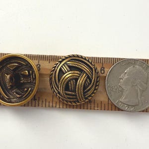 May include: Two antique brass buttons with intricate designs, next to a quarter coin for size comparison. One button has a detailed back, the other features a woven pattern. The quarter is dated 1972.