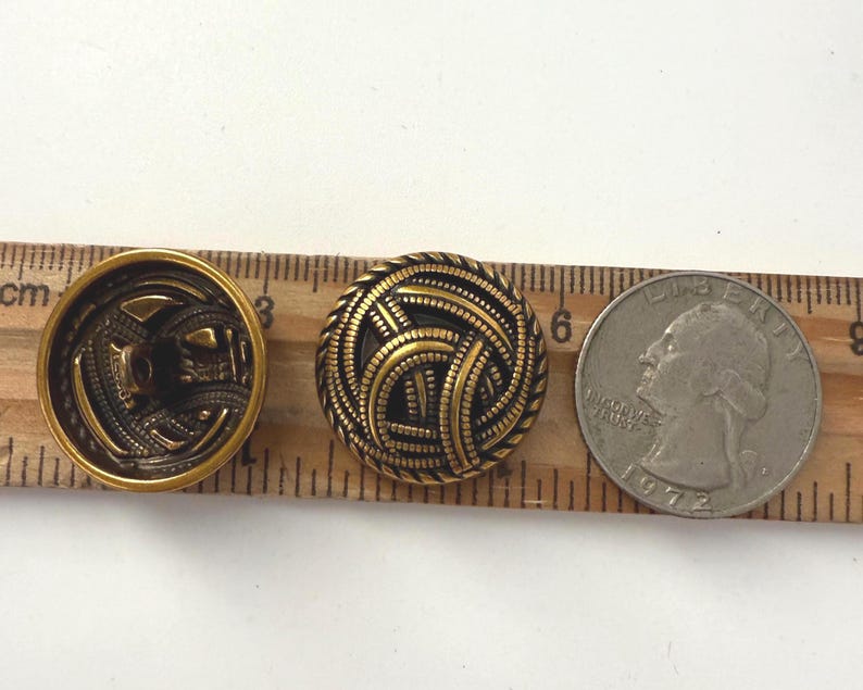 May include: Two antique brass buttons with intricate designs, next to a quarter coin for size comparison. One button has a detailed back, the other features a woven pattern. The quarter is dated 1972.