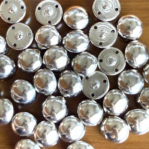 Puede incluir: Una colecci&oacute;n de botones plateados brillantes en forma de c&uacute;pula. Los botones tienen una superficie lisa y reflectante y una peque&ntilde;a base circular con agujeros para coser. Los botones est&aacute;n dispersos sobre una superficie de madera.