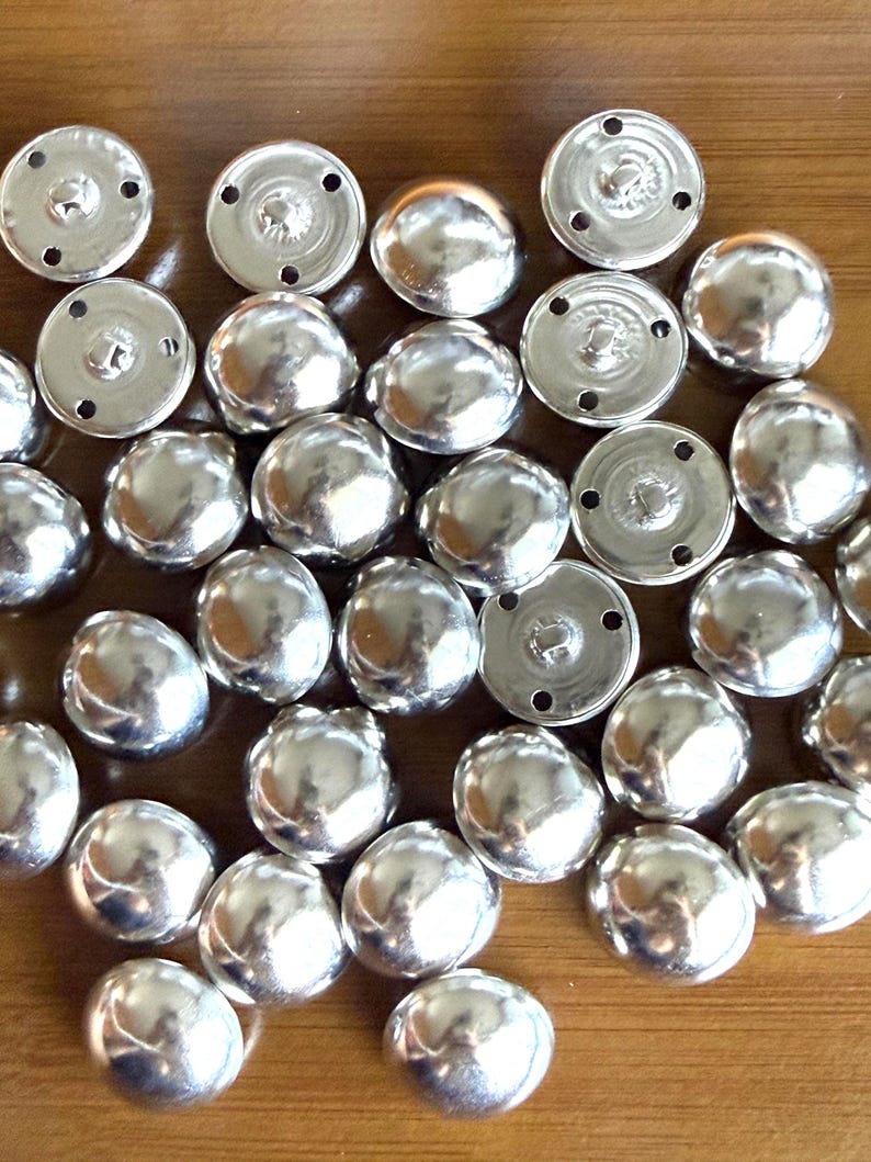 Puede incluir: Una colecci&oacute;n de botones plateados brillantes en forma de c&uacute;pula. Los botones tienen una superficie lisa y reflectante y una peque&ntilde;a base circular con agujeros para coser. Los botones est&aacute;n dispersos sobre una superficie de madera.