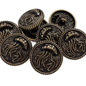 Puede incluir: Colección de botones antiguos de color latón. Cada botón presenta un relieve detallado de una medusa con tentáculos fluidos, rodeada por un borde en forma de cuerda. Los botones son redondos y tienen una estética vintage.