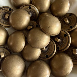 Peut inclure: Un ensemble de boutons antiques de couleur laiton. Les boutons ont un dessus arrondi et bombé et un dos plat avec une tige pour la couture. Les boutons sont de différentes tailles et sont dispersés dans un bol blanc.