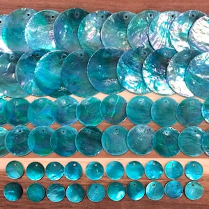 Puede incluir: Una colección de discos circulares de concha de abulón en color verde azulado iridiscente dispuestos en filas. Cada disco presenta una superficie lisa y pulida con variaciones de color naturales y un pequeño orificio cerca del borde.