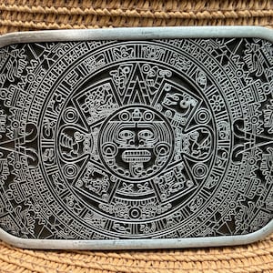 May include: A rectangular silver-colored belt buckle featuring a detailed, engraved design of the Mayan calendar. The intricate pattern includes various symbols and a central face, set against a dark background.